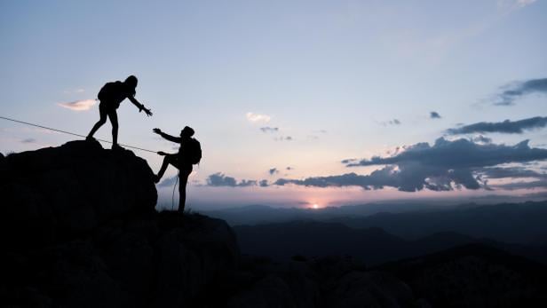 Zwei Wanderer helfen sich beim Aufstieg auf einen Felsen im Sonnenuntergang.
