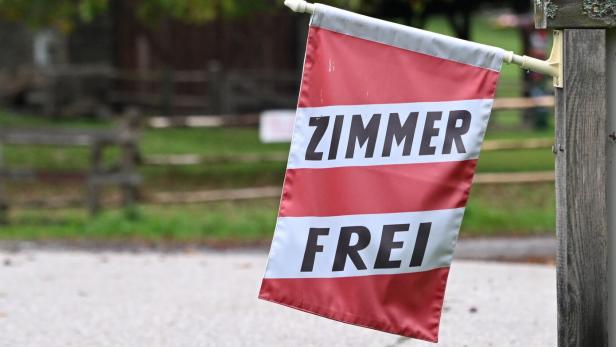 Eine rot-weiße Fahne mit der Aufschrift „Zimmer frei“ hängt an einem Holzpfosten am Straßenrand.