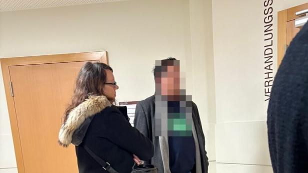 Zwei Personen stehen in einem Flur vor einer Tür mit der Aufschrift „Verhandlungssaal“.