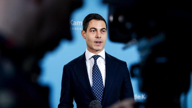 Ein Mann im Anzug spricht vor Mikrofonen und Kameras vor einem hellblauen Hintergrund.