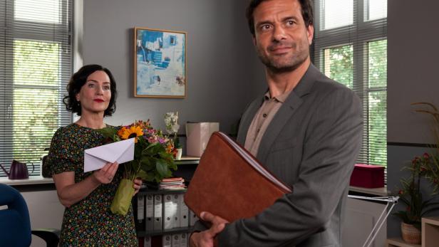 Eine Frau (Marisa Burger/ Frau Stockl) hält einen Blumenstrauß und einen Umschlag, ein Mann im Anzug steht mit einer Ledermappe daneben.