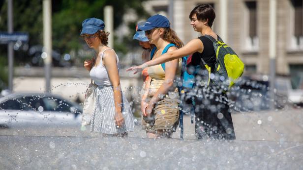 Drei junge Frauen stehen an einem sonnigen Tag an einem sprudelnden Brunnen und schauen auf das Wasser.
