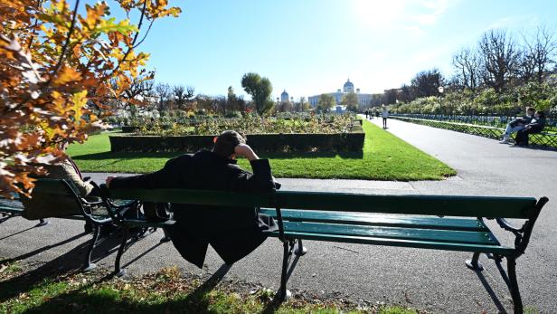 Menschen sitzen auf Bänken in einem sonnigen Park mit Blumenbeeten und einem großen Gebäude im Hintergrund.