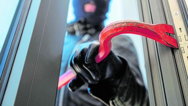 Eine vermummte Person mit Handschuhen hebelt mit einem roten Brecheisen ein Fenster auf.