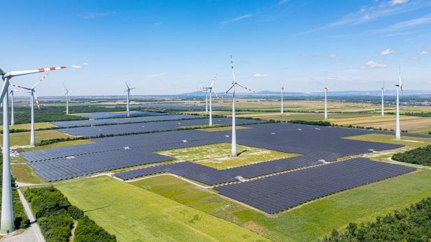 Mehrere Windräder und große Solarfelder stehen auf einer weitläufigen grünen Landschaft unter blauem Himmel.