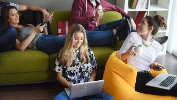 Eine Gruppe junger Leute entspannt mit Laptops, Tablets und Getränken auf einem grünen Sofa und einem gelben Sitzsack.
