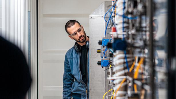 Ein Mann mit Schutzbrille betrachtet eine komplexe technische Anlage mit vielen Kabeln und Ventilen.