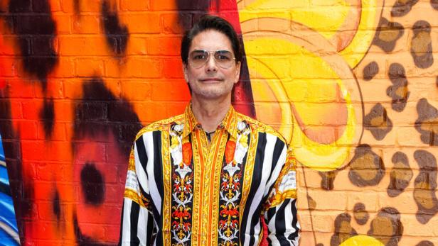 Ein Mann mit gemustertem Hemd und schwarzer Hose steht vor einer bunt bemalten Wand.