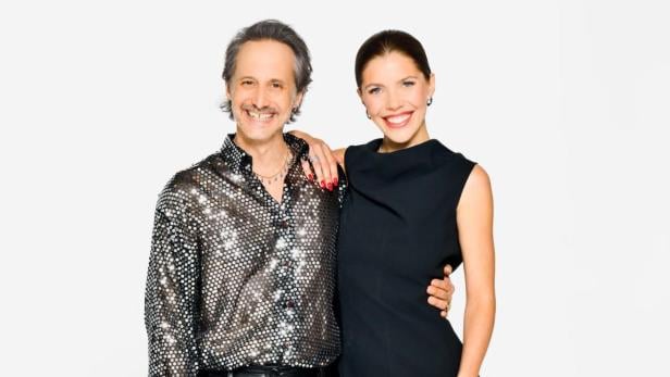 Ein Mann in Glitzerhemd und Lederhose steht neben einer Frau im schwarzen Kleid und silbernen Stiefeln.