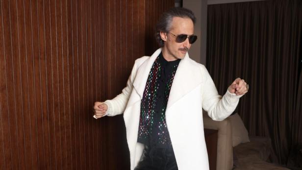 Ein Mann mit Sonnenbrille trägt weißen Mantel und schwarze Lederhose und posiert tanzend in einem modernen Raum.