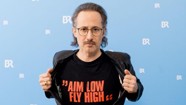Ein Mann mit Brille und Lederjacke zeigt ein T-Shirt mit dem Aufdruck "AIM LOW FLY HIGH".