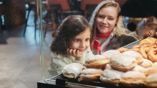 Eine erwachsene Frau kniet neben einem kleinen Mädchen, das in eine Vitrine mit Krapfen hineinschaut.