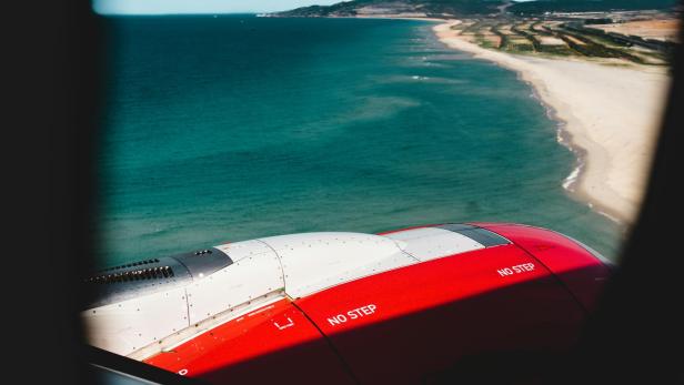 Blick aus einem Flugzeugfenster auf die Tragfläche mit der Aufschrift 'NO STEP' und eine Küstenlinie mit Strand und Meer im Hintergrund.