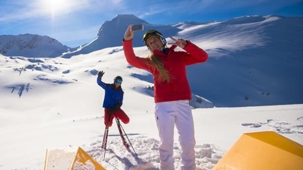 Eine Frau macht ein Selfie im Schnee, während ein Mann im Hintergrund winkt.