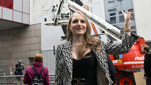 Eine Frau steht vor einer bemalten Hauswand mit der Aufschrift „Lugner City“ und zeigt auf das Wandbild.