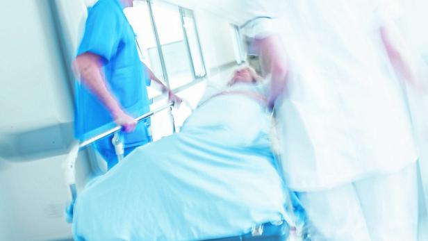 Zwei medizinische Fachkräfte schieben eine liegende Person auf einer Trage eilig durch einen Krankenhausflur.