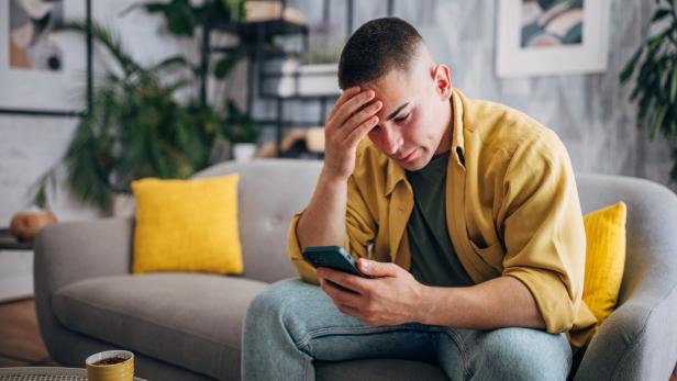 Ein Mann sitzt besorgt auf einem Sofa und schaut auf sein Handy.
