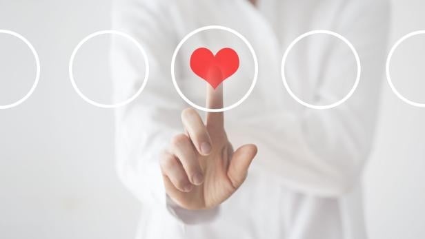 Eine Hand tippt auf ein rotes Herzsymbol, das in einem weißen Kreis auf einem transparenten Interface erscheint.