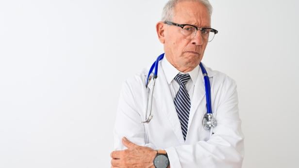 Ein älterer Arzt mit Brille, Stethoskop und Krawatte blickt nachdenklich zur Seite.