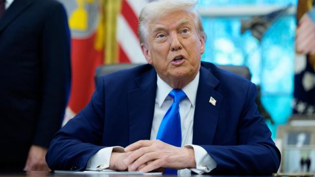 Donald Trump sitzt in einem blauen Anzug mit blauer Krawatte an einem Tisch und spricht.