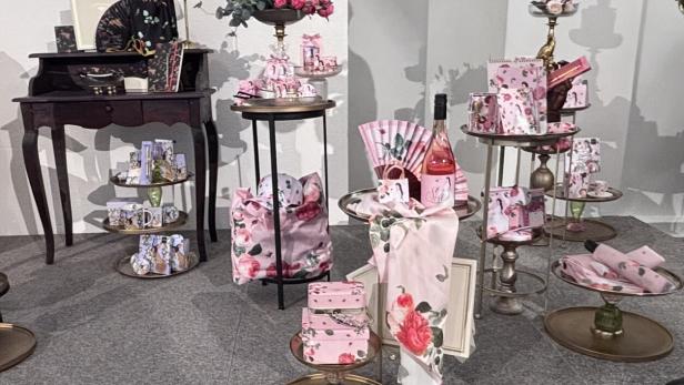 Verschiedene rosa und floral verzierte Geschenkartikel, Taschen, Schachteln und eine Flasche Roséwein sind dekorativ arrangiert.