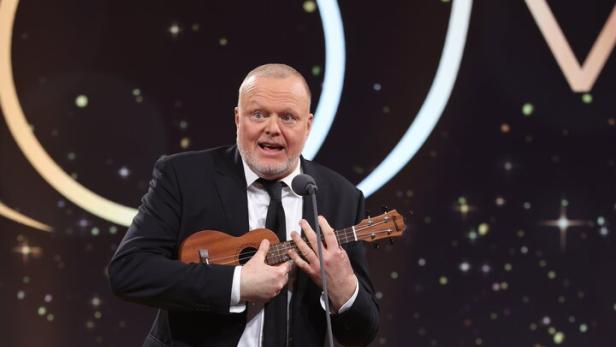 Ein Mann - Stefan Raab -  im Anzug spielt auf einer Bühne eine Ukulele und steht vor einem Mikrofon, im Hintergrund leuchten Lichter.