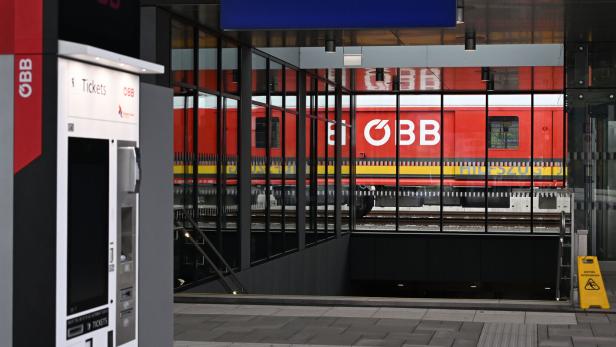Ein roter ÖBB-Zug steht hinter Glaswänden, daneben ein Fahrkartenautomat und ein Wegweiser zu den Zügen.