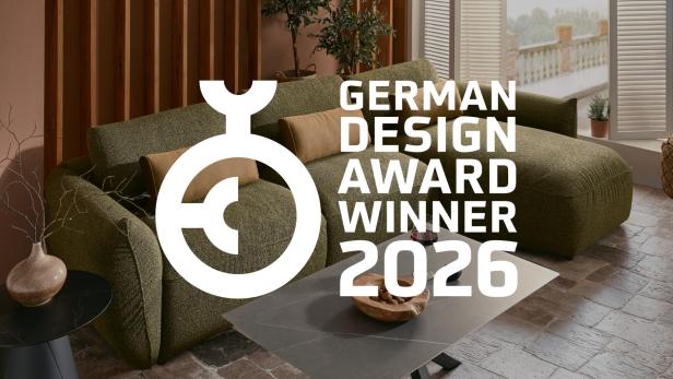 Wohnzimmerszene mit grünem Sofa, schwarzem Couchtisch und großem Fenster im Hintergrund, darüber das Logo und der Schriftzug 'GERMAN DESIGN AWARD WINNER 2026'.