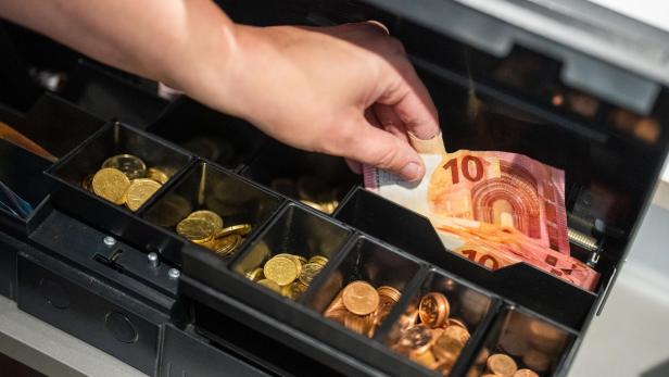 Eine Hand legt einen Zehn-Euro-Schein in eine geöffnete Ladenkasse mit Münzfächern.