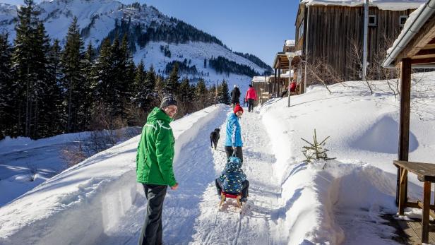 Mehrere Menschen gehen bei Sonnenschein auf einem verschneiten Weg neben Holzhäusern, umgeben von Bergen und Tannen.