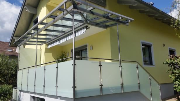 Balkon mit Edelstahlgeländer und Glasüberdachung an einem gelb gestrichenen Wohnhaus.