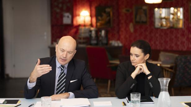 Zwei Personen im Anzug sitzen an einem Tisch mit Papieren und Wasser und führen ein Gespräch.