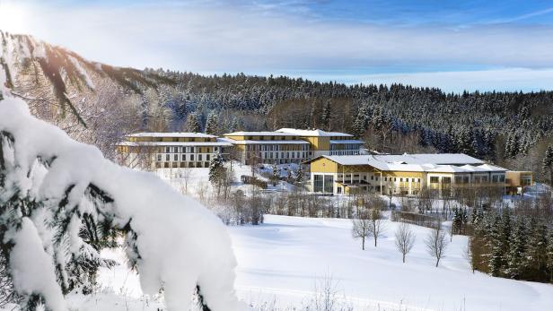 Eine Hotelanlage liegt verschneit vor einem dunklen Wald unter blauem Himmel.