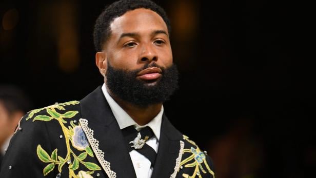 Ein Mann mit Vollbart trägt einen schwarzen Anzug mit floralen Stickereien und Schmuckdetails.