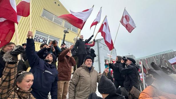 Menschenmenge mit rot-weißen Flaggen steht draußen vor Gebäuden, einige fotografieren, andere halten Fahnen hoch.