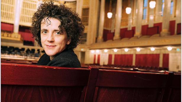 Eine Person mit lockigen Haaren - Olga Neuwirth - sitzt in einem eleganten Konzertsaal auf einer roten Holzbank und blickt lächelnd zurück.