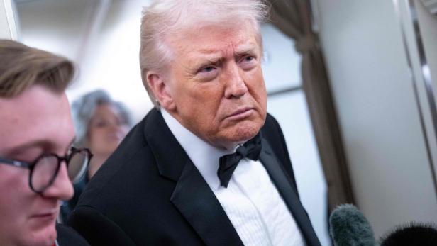 Donald Trump im Smoking, mit ernstem Blick, neben einer Person mit Brille.