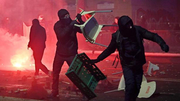 Vermummte Personen werfen Gegenstände auf einer Straße mit Rauch und Trümmern im Hintergrund.