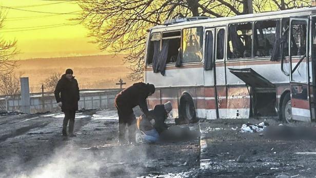 Ein zerstörter Bus steht vor einer Friedhofsmauer, Helfer kümmern sich um Verletzte.