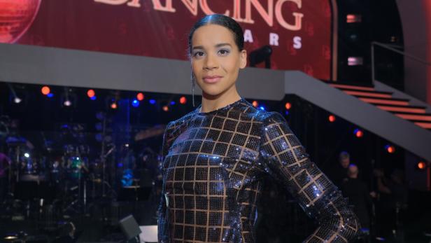 Eine Frau mit streng zurückgebundenen Haaren trägt ein glitzerndes, kariertes Kleid auf einer Bühne mit Lichtern.