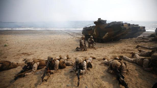 Soldaten liegen am Strand mit Gewehren, während ein amphibisches Fahrzeug im Hintergrund steht.
