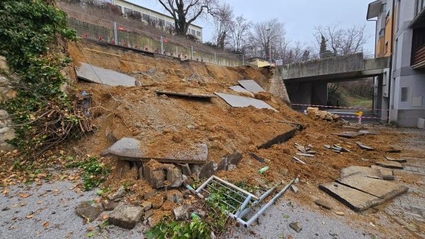 Eingestürzte Stützmauer mit Erdrutsch und Absperrungen im Bereich eines Wohngebäudes.