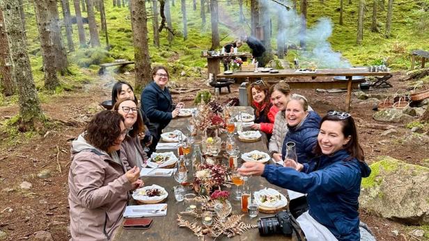 Eine Gruppe von Menschen sitzt an einem rustikalen Holztisch im schwedischen Wald und genießt gemeinsam ein Essen.