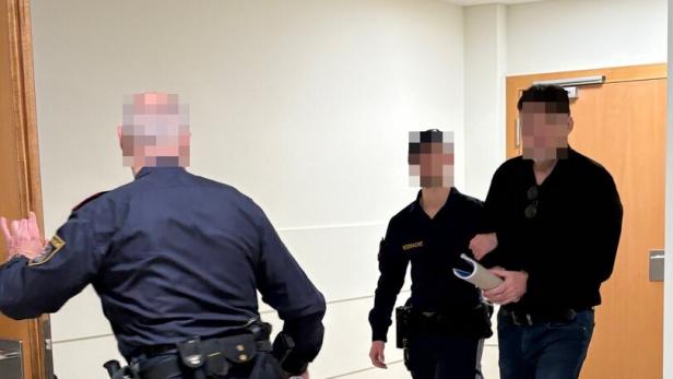 Zwei Polizisten führen einen Mann mit gefesselten Händen durch einen Flur zu einer Tür.