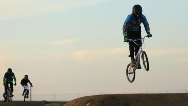 Drei BMX-Fahrer mit Helmen fahren und springen auf einer BMX-Strecke bei Tageslicht.