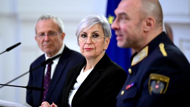 Drei Personen, darunter eine Frau mit Brille und ein Mann in Uniform, sitzen vor EU- und Landesflaggen.