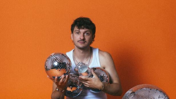 Der Musiker Rian sitzt vor orangefarbenem Hintergrund und hält mehrere Discokugeln verschiedener Größen in den Händen und auf dem Boden.