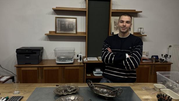 Ein junger Mann steht mit verschränkten Armen hinter einem Schreibtisch mit Silbermünzen und Schalen in einem Büro.