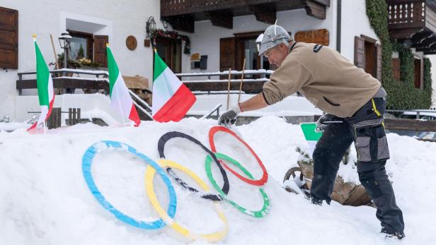 Ein Mann dekoriert für die olympischen Winterspiele 2026 im Schnee olympische Ringe, daneben stehen italienische Flaggen.