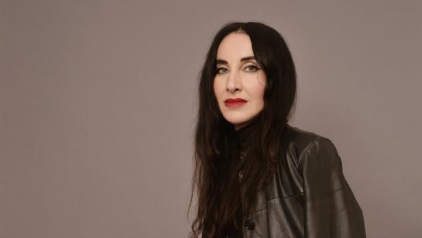 Frau -Mirna Funk -  mit langen dunklen Haaren trägt schwarze Lederjacke und schwarze Hose mit weißem Streifen, sitzt vor grauem Hintergrund.